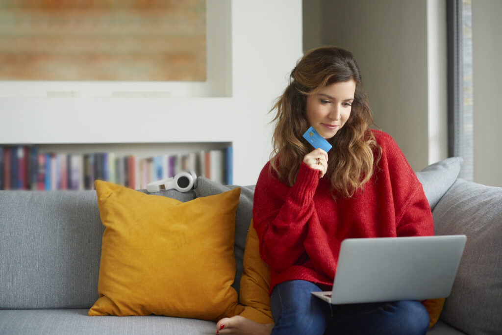 Una mujer hermosa que usa una tarjeta de crédito y una computadora portátil mientras busca el mejor programa de fidelización en su cómodo sofá.