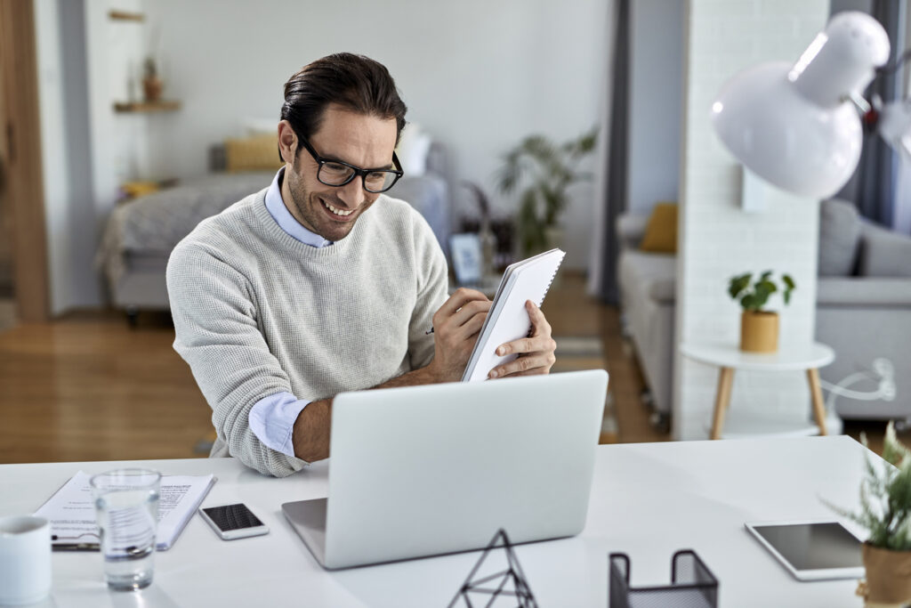 El hombre con suéter blanco y anteojos está sentado frente a la computadora y lleva un cuaderno. Escribe en el cuaderno mientras sonríe a la computadora.