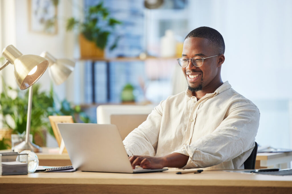 Un hombre negro con anteojos y una camisa beige está sentado en una mesa trabajando en su computadora. Está sonriendo porque estaba feliz de descubrir el programa de lealtad de American Airlines y sus beneficios