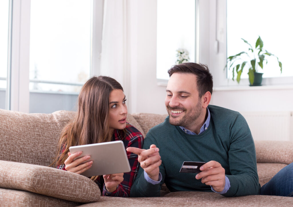 Un hombre blanco con cabello castaño y un suéter verde está acostado en el sofá al lado de su esposa, blanca, con cabello castaño lacio, vistiendo una camisa roja a cuadros. Sostiene una tarjeta de crédito mientras mira a la mujer con la tableta y le explica cómo funciona credito aerolineas argentinas