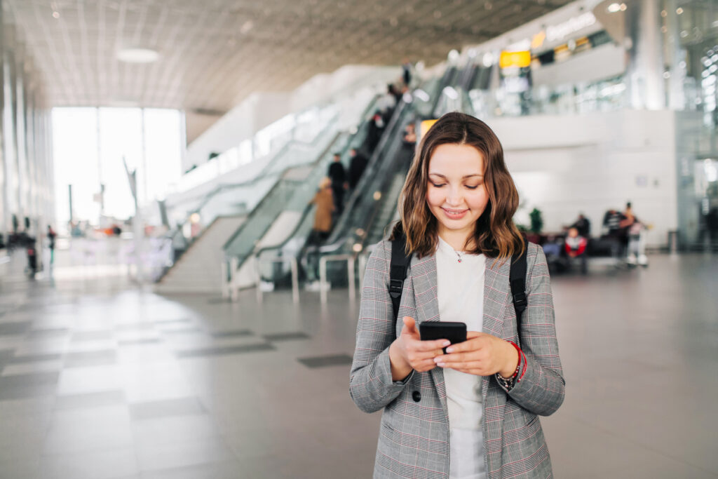 Una mujer blanca, de pelo lacio corto castaño claro, viste una blusa blanca, un blazer gris claro y una mochila negra a la espalda. Está de pie en un aeropuerto jugando con su teléfono celular y sonriendo mientras espera su viaje. Consiguió los mejores precios gracias al programa de millas smiles