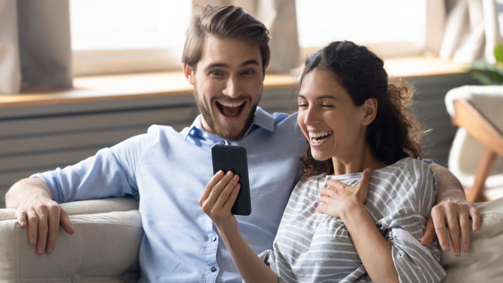 Una mujer morena con cabello castaño claro ondulado lleva una blusa gris con rayas blancas. Ella está sentada junto a su esposo, un hombre blanco con cabello rubio oscuro corto, barba rubia oscura y una camisa de vestir azul claro. Están sentados en un sofá, mientras la mujer está en su celular, están sonriendo felices porque lograron acumular mil millas rápidamente con las propinas de MercoMillas